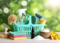 Bright turquoise caddy filled with household cleaning essentials including spray bottle, microfiber cloths, gloves and natural brushes on wooden table against green bokeh background