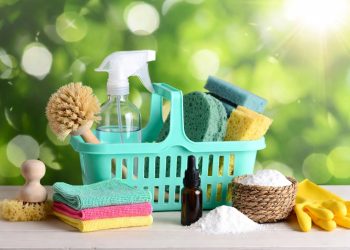 Bright turquoise caddy filled with household cleaning essentials including spray bottle, microfiber cloths, gloves and natural brushes on wooden table against green bokeh background