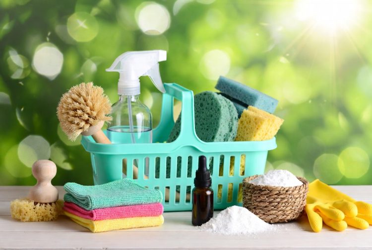 Bright turquoise caddy filled with household cleaning essentials including spray bottle, microfiber cloths, gloves and natural brushes on wooden table against green bokeh background
