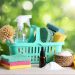Bright turquoise caddy filled with household cleaning essentials including spray bottle, microfiber cloths, gloves and natural brushes on wooden table against green bokeh background