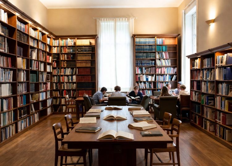Cozy library reading room with long wooden tables, bookshelves, and people studying, highlighting benefits of public library cards including free streaming services like Hoopla, Libby, Kanopy and LinkedIn Learning