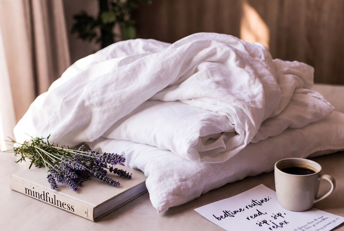 Crisp neatly folded linen sheets with lavender and self-care items on wooden surface, cozy bedroom essentials flat lay aesthetic
