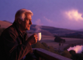 Elderly white-haired grandpa sitting on a porch at twilight, peacefully drinking coffee while looking out at a beautiful purplish sunset over scenic hills and lake, calm and nostalgic evening scene