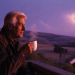 Elderly white-haired grandpa sitting on a porch at twilight, peacefully drinking coffee while looking out at a beautiful purplish sunset over scenic hills and lake, calm and nostalgic evening scene