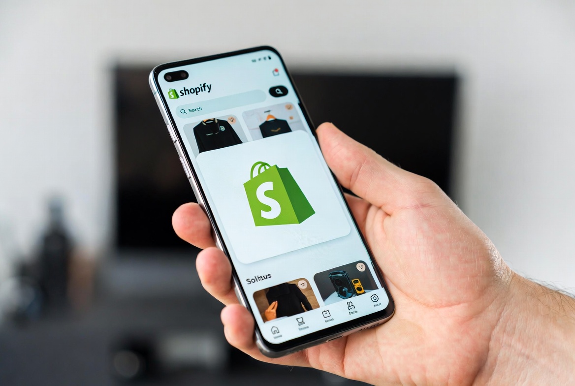 Hand holding a smartphone displaying the Shopify mobile app with green shopping bag logo and product listings, e-commerce store interface