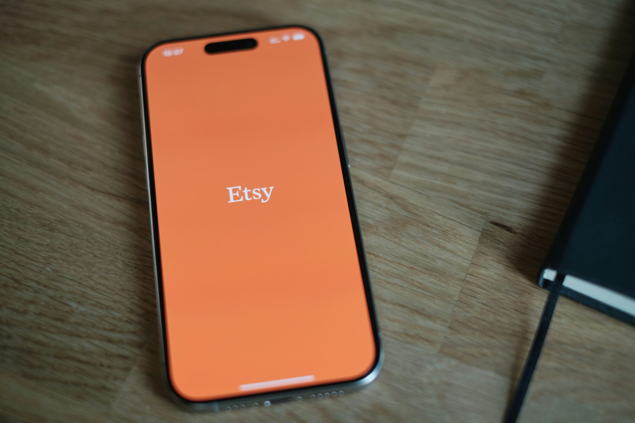 Modern smartphone lying on wooden table showing the orange Etsy app login screen with white Etsy logo, minimalist flat lay composition