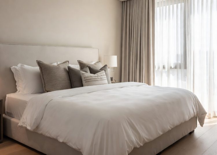 Neatly made bed with white linens and grey pillows in a bright minimalist bedroom, correct way to put on bed sheets for a perfect hotel-style finish