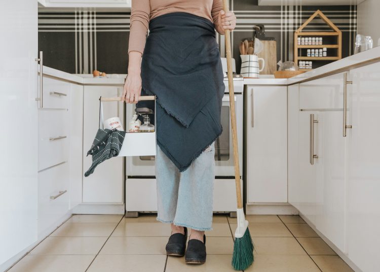 Person wearing apron holding cleaning caddy and broom in a modern kitchen, preparing for a deep clean before leaving on vacation
