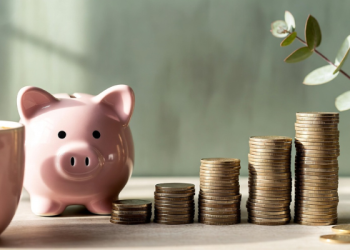 Pink piggy bank with four ascending stacks of gold coins on wooden desk against soft sage green wall, cozy aesthetic savings and financial growth flat lay with coffee mug