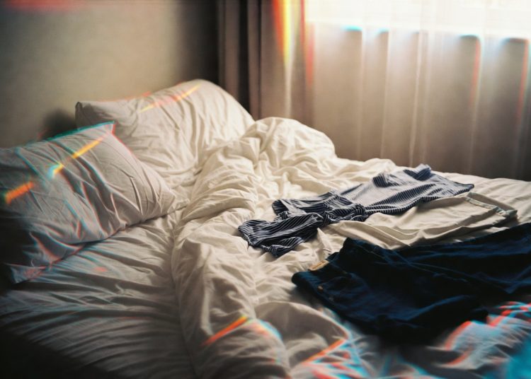 Unmade bed with rumpled white sheets and clothes scattered on top in a sunlit bedroom, classic grandma’s advice to make your bed every morning for a better start to the day