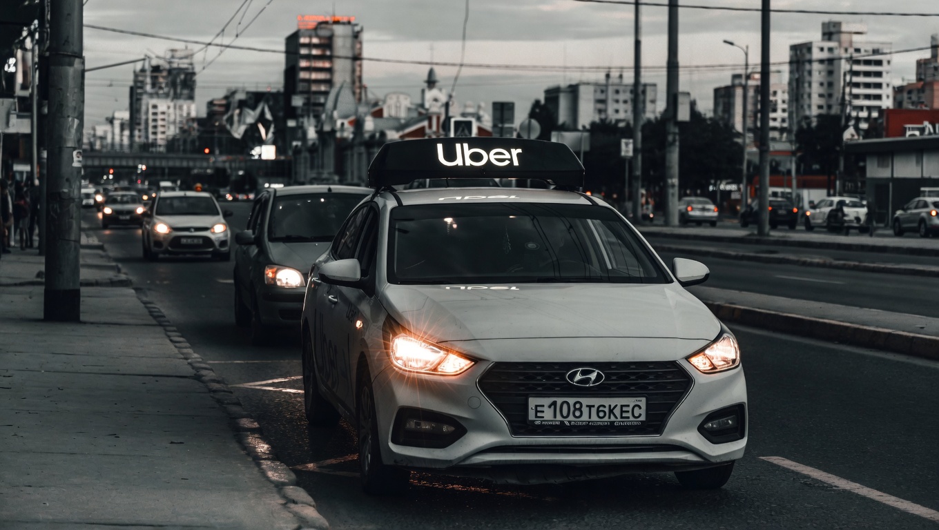 White Hyundai Uber taxi with illuminated roof sign driving on a busy city street at dusk, headlights on, urban background with buildings and traffic