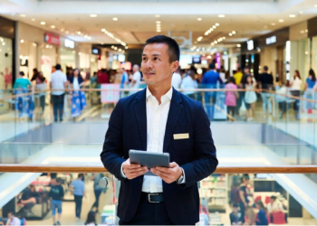 Confident retail store manager standing in a busy shopping mall, holding a tablet while overseeing operations, career growth in retail management