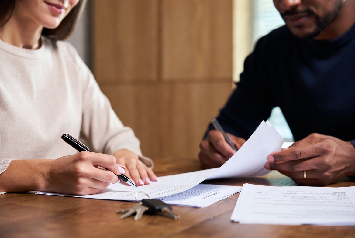 Couple signing real estate or rental agreement documents with house keys on wooden table during home purchase or lease process