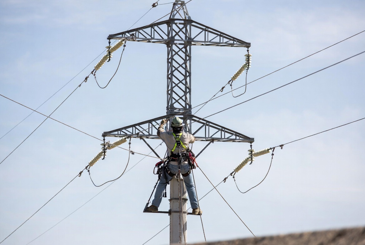 Powerline technician in safety harness working high on an electrical transmission tower against a clear blue sky