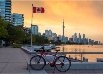 Red bicycle parked on Toronto waterfront promenade with Canadian flag waving at sunset, CN Tower and city skyline in background