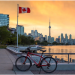 Red bicycle parked on Toronto waterfront promenade with Canadian flag waving at sunset, CN Tower and city skyline in background